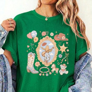 May include: A vibrant green t-shirt featuring a central graphic print of a person playing a guitar inside an ornate gold frame, surrounded by illustrations of a cowboy boot, a hat, a teddy bear, a rocking horse, balloons, and flowers.