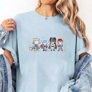 May include: A light blue t-shirt featuring a watercolour-style graphic of four chibi-proportioned superheroes standing in a row. The characters include Thor with a hammer, Captain America with a shield, Batman, and Spider-Man, accented by colourful paint splatters.