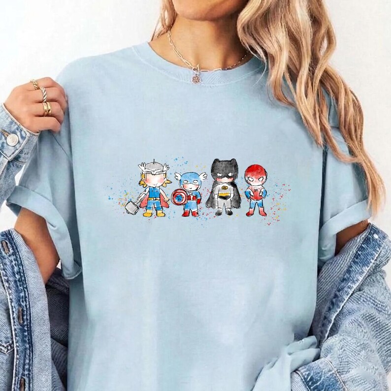 May include: A light blue t-shirt featuring a watercolour-style graphic of four chibi-proportioned superheroes standing in a row. The characters include Thor with a hammer, Captain America with a shield, Batman, and Spider-Man, accented by colourful paint splatters.