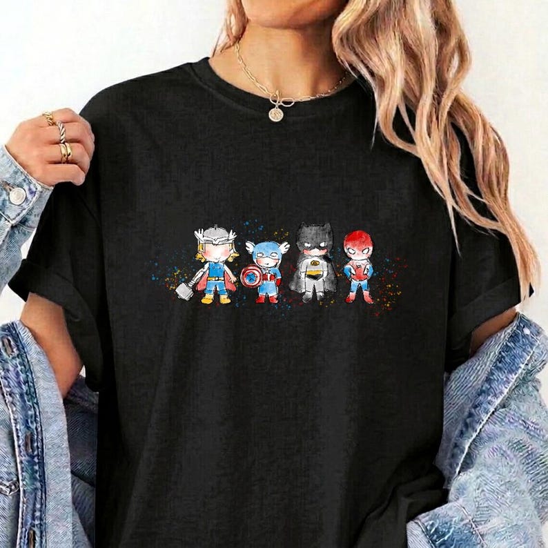 May include: A black short-sleeved t-shirt featuring a graphic print of four stylised, chibi-style superhero characters standing in a row, accented by colourful paint splatters in the background, with a crew neckline and a relaxed fit.