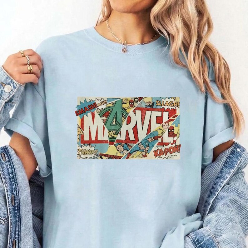 Puede incluir: Una camiseta de manga corta de color azul claro con un estampado gr&aacute;fico rectangular central. El dise&ntilde;o muestra la palabra MARVEL en letras blancas grandes sobre un collage de paneles de c&oacute;mics antiguos con textos de acci&oacute;n como SMASH y KA-POW.