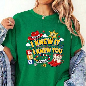 Puede incluir: Una camiseta verde con un estampado gr&aacute;fico que incluye un sombrero de vaquero rojo, una estrella de sheriff amarilla con el texto YEP, I KNEW IT, y el texto I KNEW IT I KNEW YOU. Incluye ilustraciones de bloques de letras, una pulsera y botas rojas.