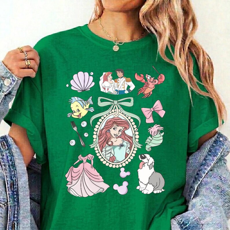 May include: A green short-sleeve t-shirt featuring a central oval frame containing a cartoon character with long red hair. Surrounding the frame are various illustrations including a seashell, a fork, a pink bow, a crab, and a white dog.