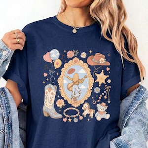May include: A navy blue t-shirt featuring a central ornate gold frame containing an illustration of a person playing a guitar. Surrounding the frame are graphics of a cowboy boot, a hat, a star, a teddy bear, a rocking horse, and balloons.
