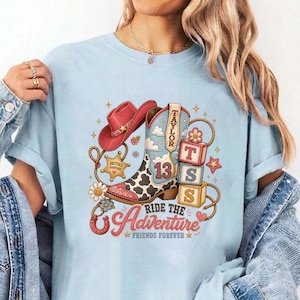K&ouml;nnte beinhalten: Ein hellblaues T-Shirt mit einem Grafikdruck, der einen rosa Cowboyhut, einen Cowboystiefel mit Kuhmuster und der Zahl 13 sowie drei gestapelte Bl&ouml;cke mit den Buchstaben T, S und S zeigt. Das Design enth&auml;lt den Text "TAYLOR", "Ride the Adventure" und "FRIENDS FOREVER".