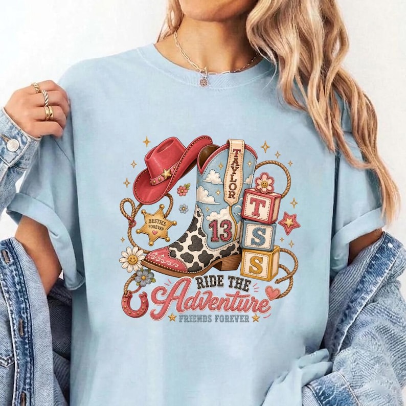 K&ouml;nnte beinhalten: Ein hellblaues T-Shirt mit einem Grafikdruck, der einen rosa Cowboyhut, einen Cowboystiefel mit Kuhmuster und der Zahl 13 sowie drei gestapelte Bl&ouml;cke mit den Buchstaben T, S und S zeigt. Das Design enth&auml;lt den Text "TAYLOR", "Ride the Adventure" und "FRIENDS FOREVER".
