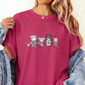 May include: A magenta short-sleeved t-shirt featuring a graphic print of four stylised superhero characters standing in a row. The characters include figures resembling Thor, Captain America, Batman, and Spider-Man in a small, cartoon-like design.