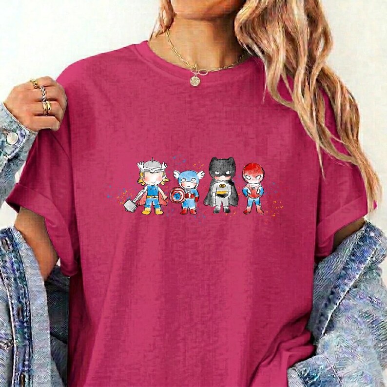 May include: A magenta short-sleeved t-shirt featuring a graphic print of four stylised superhero characters standing in a row. The characters include figures resembling Thor, Captain America, Batman, and Spider-Man in a small, cartoon-like design.