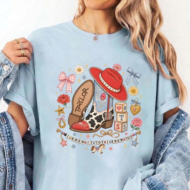 May include: A light blue t-shirt featuring a graphic print of a cow-print cowboy boot, a red cowboy hat, alphabet blocks, and floral elements. The text "TAYLOR" is on the boot, "13" on the heel, and "I KNEW IT TOY I KNEW YOU" on a banner.