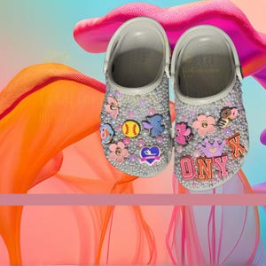 May include: A pair of gray Crocs shoes heavily embellished with faux pearls and decorative charms. Charms include flowers, a baseball, a blue cartoon character, and the name "ONYX". The shoes have a strap and are set against a colorful background.