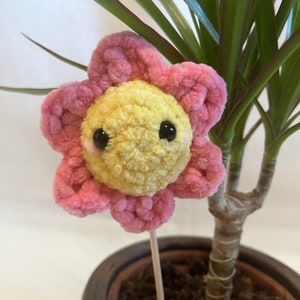 Könnte beinhalten: Eine gehäkelte Blume mit gelbem Zentrum und rosa Blütenblättern. Die Blume hat schwarze Augen und einen fröhlichen Ausdruck. Sie ist an einem Holzstab befestigt und steht in einem Topf mit einer grünen Pflanze.