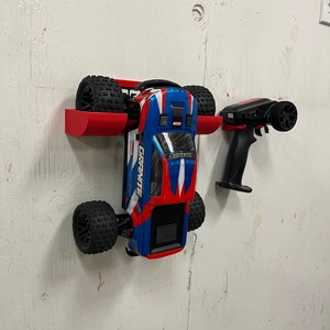 Peut inclure: Un support mural rouge pour une voiture télécommandée et une télécommande. La voiture est bleue, rouge et blanche avec des roues noires. La télécommande est noire avec un bouton rouge. Le support mural est en plastique rouge.