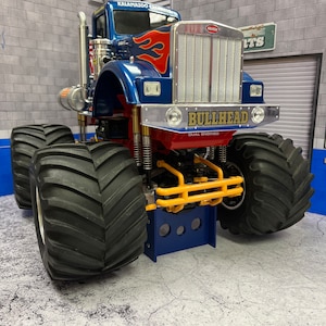 May include: A blue and red monster truck with the words "Bullhead Dual Engine" on the front. The truck is on a blue platform with a grey concrete background.