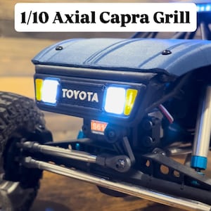 May include: A close-up of a blue 1/10 Axial Capra Grill with the Toyota logo. The grill has bright white headlights and a yellow accent. The image is of a model car.