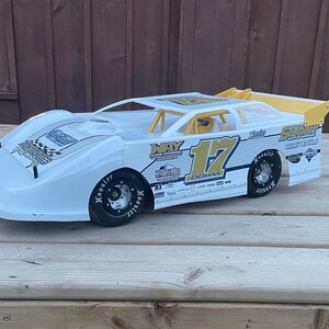 May include: White race car with yellow accents and the number 17 on the side. The car has a black spoiler and black wheels with white lettering. The car is on a wooden surface.