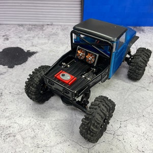 Pode incluir: Um caminh&atilde;o de brinquedo azul e preto com uma gaiola de seguran&ccedil;a preta e ventiladores vermelhos. O caminh&atilde;o tem pneus pretos grandes e est&aacute; sentado em uma superf&iacute;cie de concreto cinza.