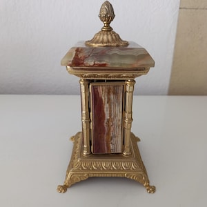May include: A rectangular clock featuring a polished gold-tone metal frame with ornate detailing, four pillars, and a base with claw feet. The clock is accented with panels of banded brown and cream onyx stone and topped with a decorative finial.
