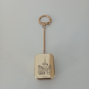 Peut inclure: Un porte-clés rectangulaire doré avec une chaîne et un anneau. Le porte-clés présente une illustration détaillée en noir et blanc d'un bâtiment avec une tour d'horloge, avec le mot "BERN" en dessous.