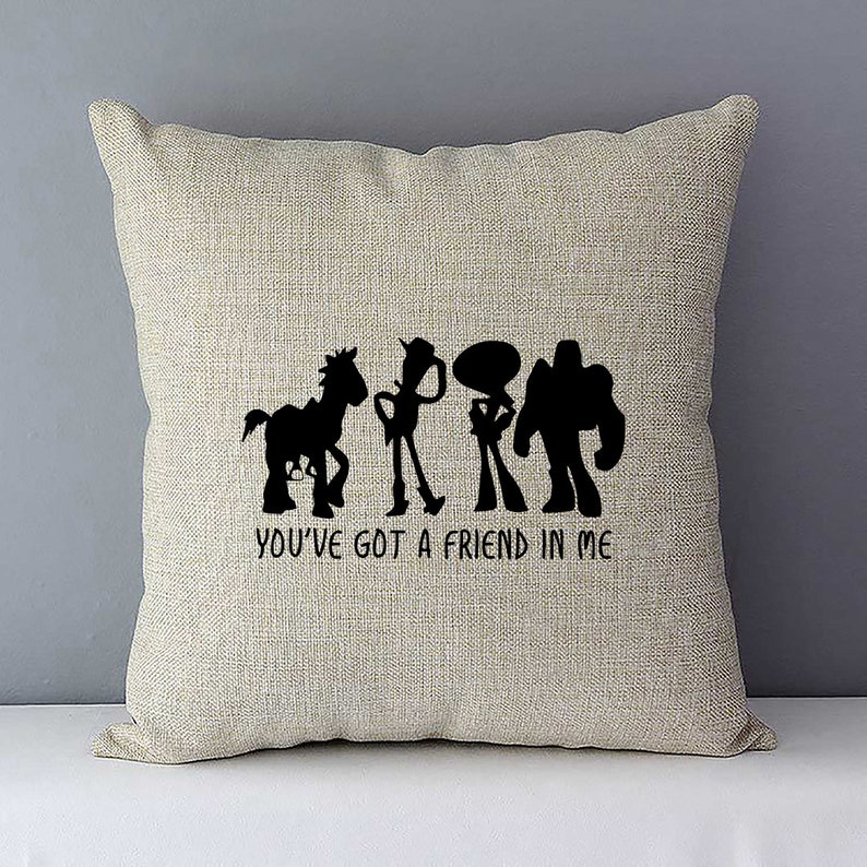 May include: A beige throw pillow with a black silhouette of the Toy Story characters Woody, Buzz Lightyear, Jessie, and Bullseye. The text "You've got a friend in me" is printed below the characters.