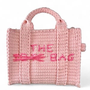 May include: A light pink crocheted tote bag with the words "THE BAG" embroidered in pink. The bag has a handle and vertical stripes. The word "SO" is crossed out with pink yarn.