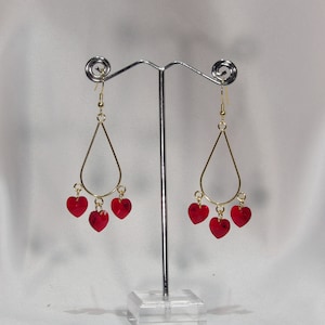 May include: Gold dangle earrings with three red heart-shaped charms hanging from each earring.