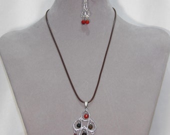 May include: A silver necklace with a pendant featuring a cluster of red and brown stones. The pendant is shaped like a teardrop and hangs from a brown cord. The necklace is paired with matching earrings.