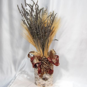 May include: A decorative arrangement featuring dried lavender, wheat, and a pinecone, presented in a birch bark vase. The arrangement is accented with a burgundy and gold ribbon. The overall theme is rustic and natural.
