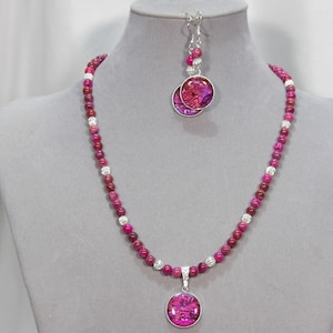 May include: A necklace and earring set displayed on a mannequin. The necklace features a strand of pink and red beads with silver accents and a matching pendant. The earrings have a similar design with a circular pendant and silver hooks.