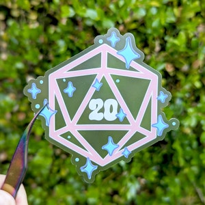 May include: A clear sticker with a pink 20-sided die with a green background and blue glitter. The die has the number 20 written on it.