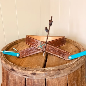 May include: A brown leather belt with floral tooling and a turquoise brand. The belt is displayed on a wooden barrel.