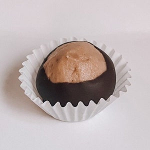 May include: A chocolate-covered confection resembling a buckeye nut, with a light brown peanut butter center. The treat sits in a white, ruffled paper cup against a white background. A sweet treat.
