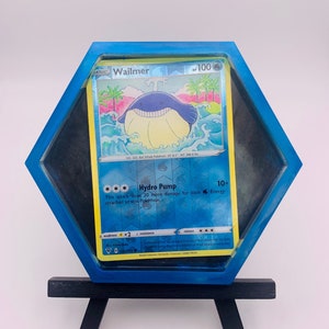 May include: A blue and black Pokemon card holder with a Wailmer Pokemon card inside. The card features a blue and white Wailmer with a yellow belly. The card holder is shaped like a hexagon and has a black easel.