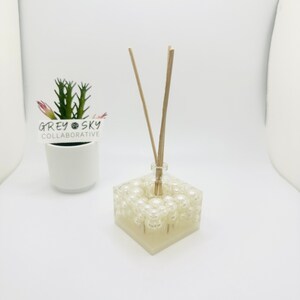 May include: A white square diffuser with a clear glass bottle and wooden sticks. The diffuser is filled with white pearl-like beads. The diffuser is sitting on a white surface.