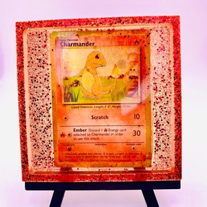 May include: A vintage Charmander Pokemon card encased in resin with red glitter. The card features an illustration of Charmander, a lizard Pokemon, with the text "Basic Pokemon Charmander" and "Lizard Pokemon. Length: 2' 0", Weight: 19 lbs."