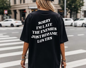 May include: Black oversized t-shirt with the text "SORRY I'M LATE THE ENEMIES JUST BECAME LOVERS" in white. The shirt is worn by a person walking on a city street. The person is wearing a black cap and carrying a black purse.