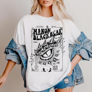 May include: White t-shirt featuring a black and white graphic of a dragon's eye and the text "MANON BLACKBEAK WORLD TOUR 13". The shirt also includes the text "THE THIRTEEN PRESENTS" and a list of names.