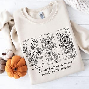 May include: A cream-colored sweatshirt featuring a floral design in black ink. The design includes three rectangular panels with flowers, moons, and stars. The text "the world will be saved and remade by the dreamers" is printed below the panels.