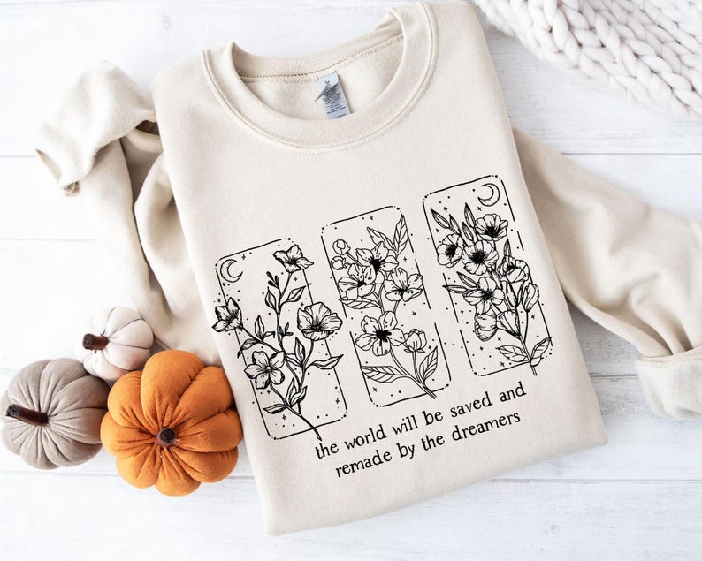 May include: A cream-colored sweatshirt featuring a floral design in black ink. The design includes three rectangular panels with flowers, moons, and stars. The text "the world will be saved and remade by the dreamers" is printed below the panels.