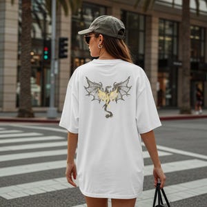 May include: White oversized t-shirt featuring a dragon graphic on the back. The dragon design has gray wings with yellow accents and a black tail. The shirt is worn by a person in a city setting.