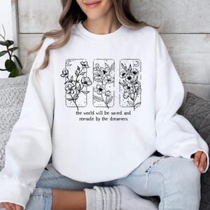 May include: White sweatshirt featuring a floral design in black ink. The design includes three rectangular panels with different floral arrangements and the text "the world will be saved and remade by the dreamers" in a simple font. The sweatshirt has long sleeves.