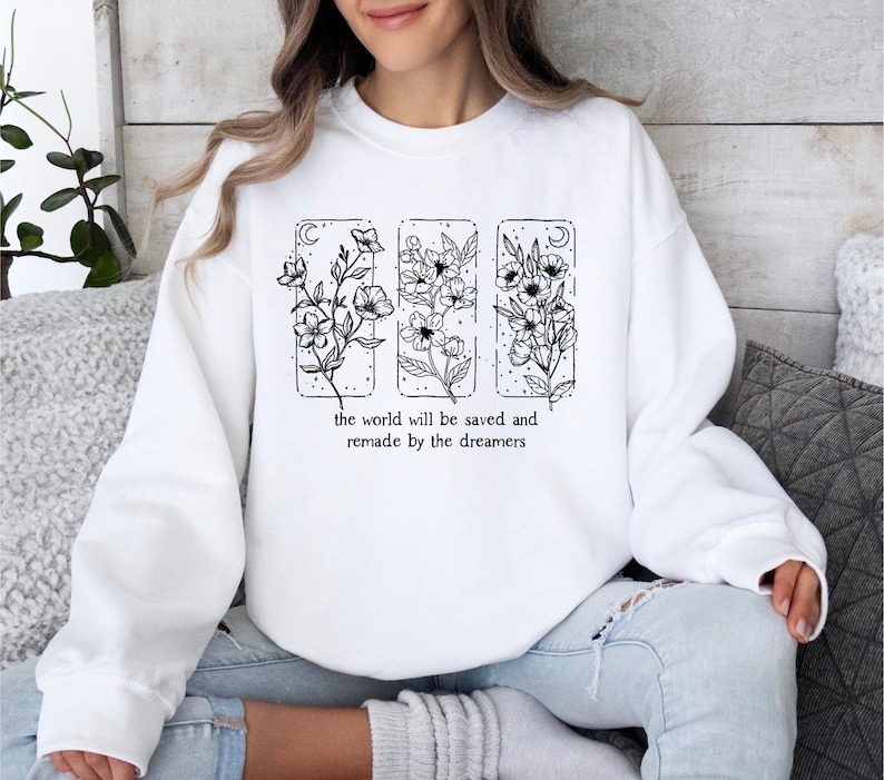 May include: White sweatshirt featuring a floral design in black ink. The design includes three rectangular panels with different floral arrangements and the text "the world will be saved and remade by the dreamers" in a simple font. The sweatshirt has long sleeves.