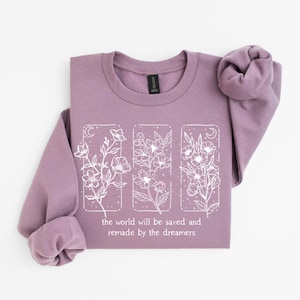 May include: A mauve-colored sweatshirt with long sleeves. The front features a white floral design within three rectangular frames, accompanied by the text: "the world will be saved and remade by the dreamers."