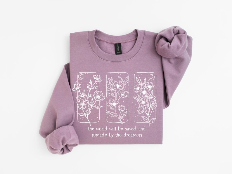 May include: A mauve-colored sweatshirt with long sleeves. The front features a white floral design within three rectangular frames, accompanied by the text: "the world will be saved and remade by the dreamers."