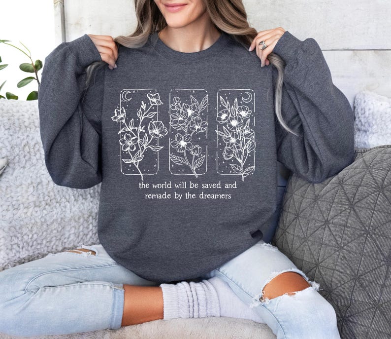 May include: Dark gray sweatshirt with white floral design and the text "the world will be saved and remade by the dreamers." The design features three vertical rectangles with floral illustrations.