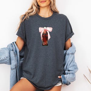 May include: Dark grey t-shirt with a watercolour illustration of a Coca-Cola bottle tied with a pink bow. The shirt is part of a casual clothing collection. The model is wearing a denim jacket.