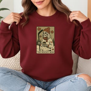 May include: A maroon sweatshirt with a graphic of a skeleton wearing a toque and holding a hockey stick and a cup of coffee. The graphic is surrounded by a red and white Canadian flag and the text "The Hockey Mom".