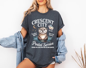 May include: Dark grey t-shirt featuring an otter graphic and the text "CRESCENT CITY Postal Service". The design includes the phrases "Friendly & Efficient" and "Same day Delivery", plus a crescent moon and stars.