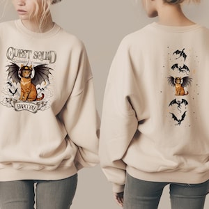 May include: A cream-coloured sweatshirt featuring a cartoon cat with bat wings and a helmet, with the text "QUEST SQUAD" and "BADCOW". The back has a similar design with the cat and bats.