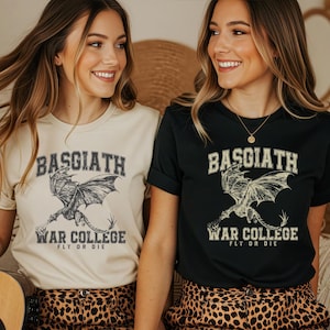 May include: Two women wearing t-shirts with a dragon graphic. One shirt is beige, the other is black. The text on the shirts reads "BASGIATH WAR COLLEGE FLY OR DIE". The dragon is detailed with wings spread.