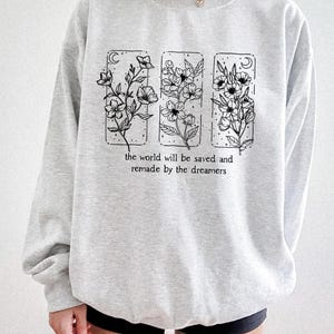 May include: A light gray sweatshirt featuring a floral design in three rectangular frames. Each frame contains a different floral arrangement with a crescent moon. The text "the world will be saved and remade by the dreamers" is printed below the frames.